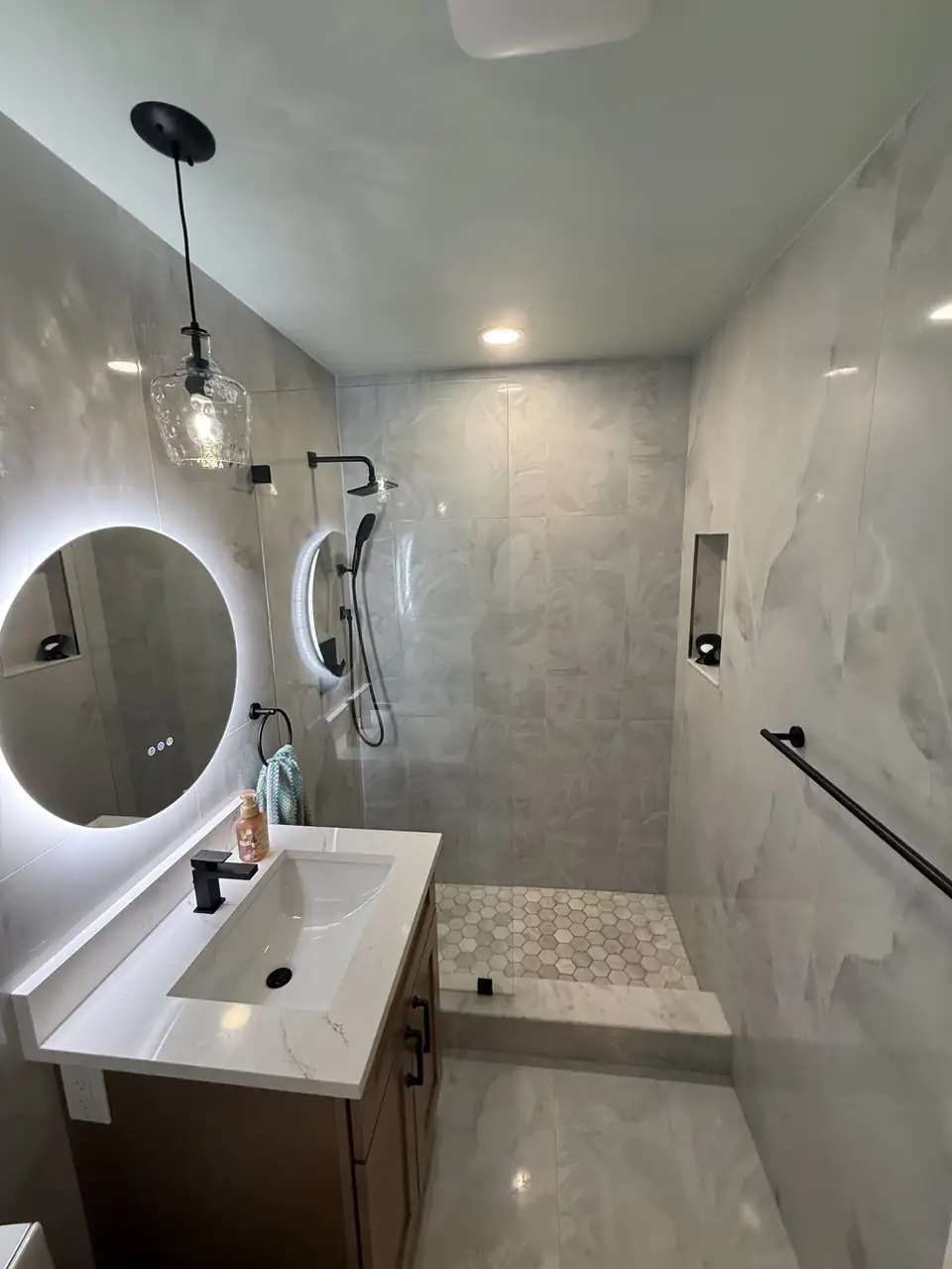 Bathroom remodeling Delray Beach FL: walk-in shower with large gray stone-look tile, mosaic floor, grab bar and a backlit round mirror over a floating vanity