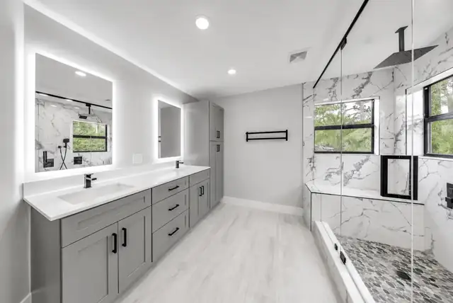 Remodeled bathroom with gray shaker double vanity, backlit mirrors and a marble-look walk-in shower with pebble floor
