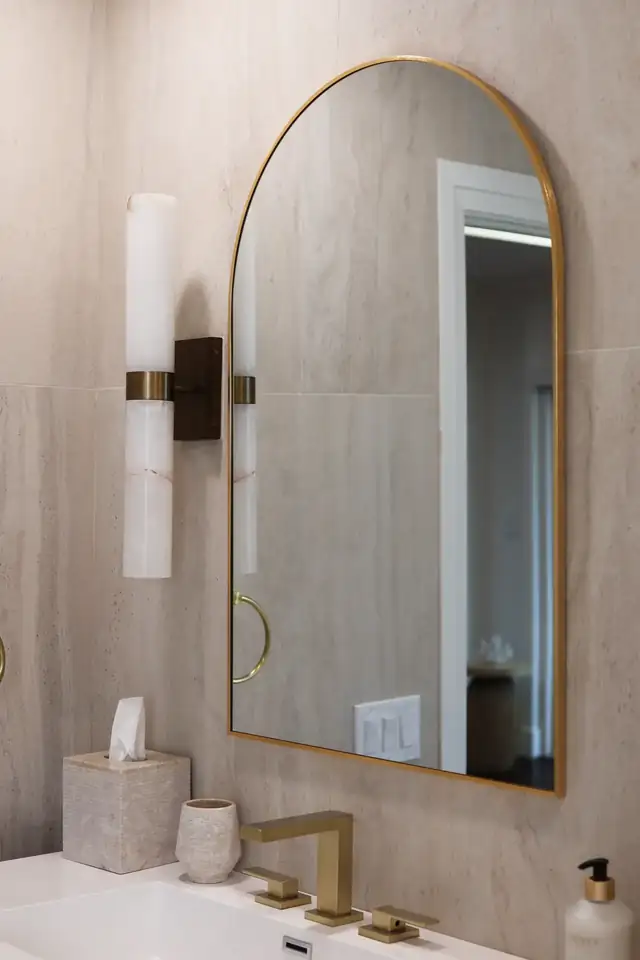 Bathroom vanity with arched brass mirror, wall sconce and beige large-format tile