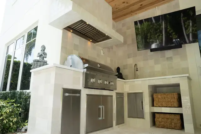 Outdoor kitchen with stainless steel cabinets, built-in grill, tile hood and mounted TV under a wood ceiling